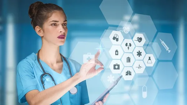 A healthcare professional in blue scrubs uses a tablet and gestures toward a transparent digital interface displaying medical icons and health data in a modern clinical setting.