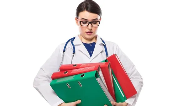 A medical professional in a white lab coat with a stethoscope around their neck struggles to carry a large stack of colorful binders, conveying a sense of being overwhelmed by paperwork. The background is plain white, emphasizing the subject and the burden. 