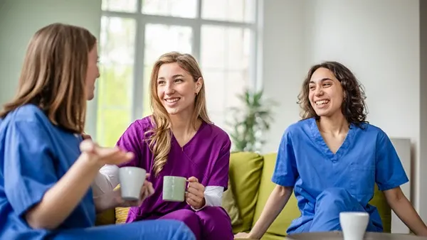 Photo of three healthcare workers in blue and purple scrubs seated together in a bright lounge area, holding mugs and conversing on a couch near a coffee table.