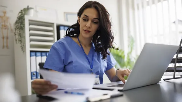 Focused female healthcare leader in blue scrubs reviews paperwork and types on a laptop, illustrating aligned healthcare operations and financial management.