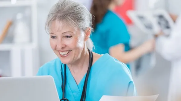 Cheerful healthcare leader reviewing digital records on a computer while completing paperwork at their desk.