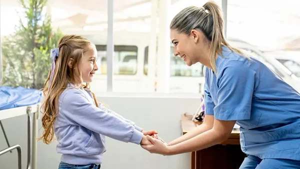 A healthcare worker in blue scrubs kneels to hold hands with a young girl student in a casual outfit, offering support in a bright school setting.