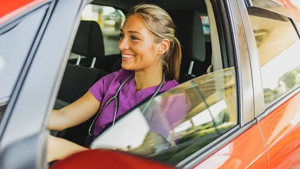 A nurse wearing scrubs and a stethoscope sits in the driver’s seat of a car, looking out the window as she prepares to travel for work.
