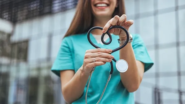 A healthcare worker in teal scrubs holds a stethoscope shaped into a heart, standing in front of a modern glass building.